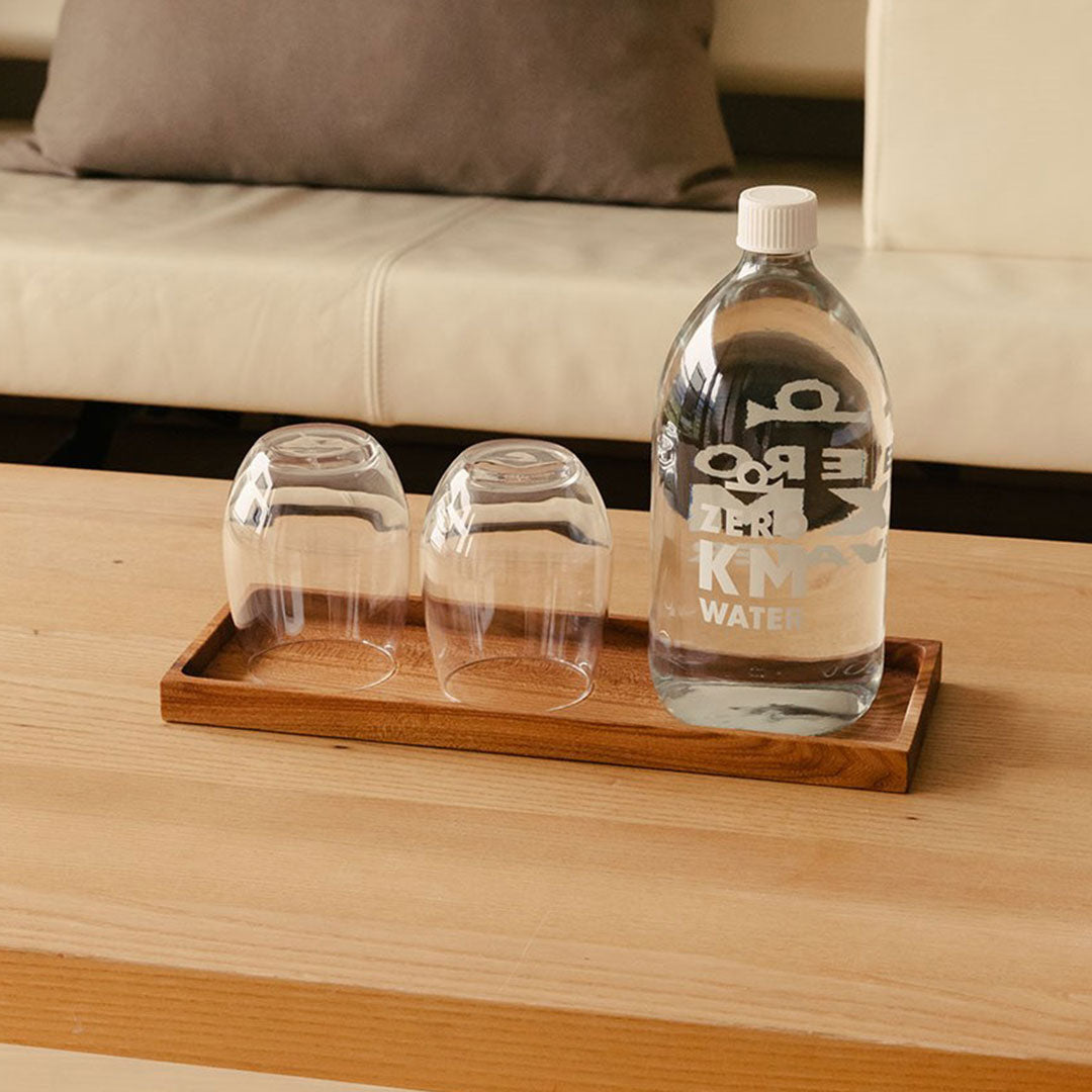 Mit Wasser gefüllte Hotel Post Bezau Glasflasche ZERO KM WATER 1 l auf dem Hotel Post Bezau Holzbrett mit zwei leeren Wassergläsern auf einem Tisch vor einem Sofa